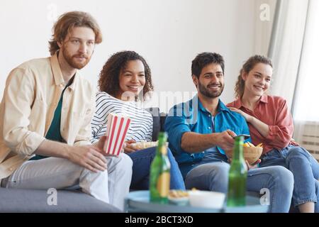 Fröhliche, multiethnische Gruppe von Freunden, die gemeinsam Fernsehen, während sie zu Hause auf einem bequemen Sofa sitzen und Snacks essen, Kopierraum Stockfoto