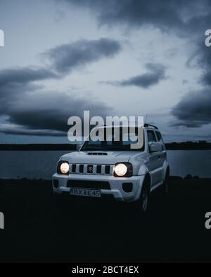 Suzuki Jimny Parkplatz neben dem See in einer bewölkten Nacht. Nachtauto mit hellen Scheinwerfern, die sich im Dunkeln nähern. Stockfoto