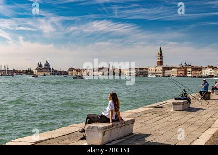 Frau auf einer Steinbank in der Sonne am Wasser in der Nähe von Arsenale mit St. Mark's Campanile und dem Dogenpalast im Hintergrund, Venedig Stockfoto