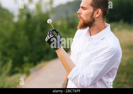 Mann hält Löwenzahn in einer bionischen Hand und bläst darauf. Seitenansicht. Stockfoto