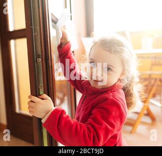 Das dreijährige Mädchen hilft bei der Haushaltsreinigung. Sie reinigt die Fenster mit einem Blatt Papier. Idee, die Zeit während der Kovid-19-Coronavirus-Quarantänezeit zu verstreichen. Stockfoto