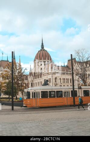 Budapest, Ungarn - 6. November 2019: Öffentliche gelbe Straßenbahn vor dem ungarischen Parlamentsgebäude. Menschen auf der Straße. Hauptstadt des öffentlichen Personennahverkehrs. Stadtverkehr. Vertikales Foto. Stockfoto