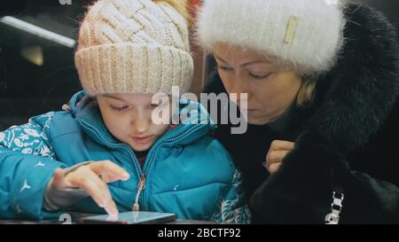 Attraktive kaukasische Mutter und Tochter mit Smartphone im Café. Fortgeschrittene kleine Kinder zeigen und erklären Mama etwas auf dem Smartphone. Stockfoto