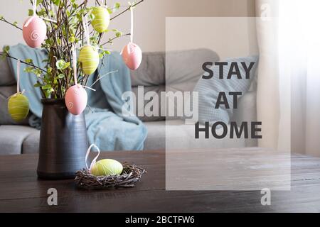 Oster-Heim-Interieur. Vase mit frischen Birkenzweigen schmückten Eier und nisten. Text - zu Hause bleiben. Selbstisolierung. COVID-19. Quarantäne. Stockfoto