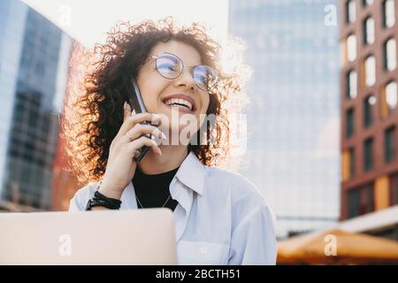 Nahaufnahme des Porträts eines kaukasischen Unternehmers mit lockigen Haaren und Brillen, die während der Arbeit mit einem Computer im Freien telefonieren Stockfoto