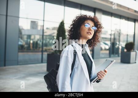 Charmante kaukasische Geschäftsfrau mit lockigen Haaren und Brillen, die auf jemanden wartet, während sie einen Laptop hält und eine Tasche trägt Stockfoto