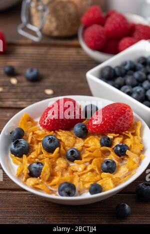 Frühstück mit Maisflocken mit Blaubeeren und Erdbeeren in weißer Schüssel auf Holztisch. Stockfoto