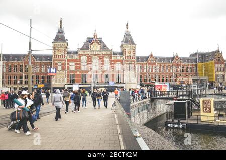 Amsterdam, Niederlande - 27. April 2019: Menschen in den Straßen der niederländischen Hauptstadt. Amsterdam Centraal, die Gebäude des Hauptbahnhofs im Hintergrund. Stadtzentrum, Kanal. Übergiebeltem Himmel. Stockfoto