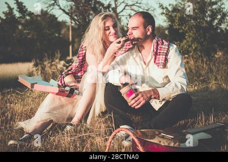 Romantisches Paar umarmt während Picknick im Sonnenuntergang Licht. Schöner bärtiger Mann mit Gitarre und attraktive junge Frau sitzen zusammen auf Gras Stockfoto