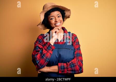 Junge afro-amerikanische afro-Bauernfrau mit geschweiften Haaren, Schürze und Hut, die mit einem Lächeln mit gekreuzten Armen und Händen auf die Kamera blickt Stockfoto