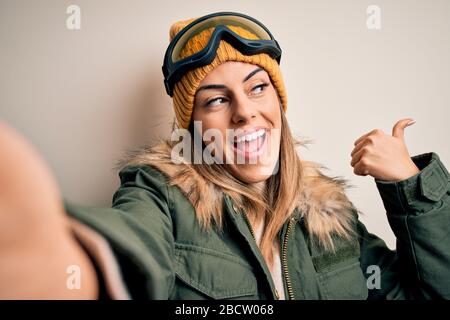 Junge Frau des Brunette-Skifahrers, die Schneekleidung und eine Skibrille trägt, macht selfie durch Kamerafunkeln und zeigt mit dem Daumen nach oben mit einem glücklichen Gesicht Stockfoto