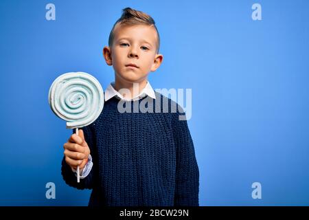 Kleines kaukasisches Kind, das süße Süßigkeiten-Lollipop über blauem isoliertem Hintergrund mit einem selbstbewussten Ausdruck für ein kluges, ernsthaftes Denken im Gesicht isst Stockfoto