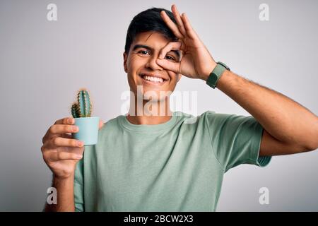 Junger, gutaussehender Mann, der den kleinen Kaktuspflanzentopf über einem isolierten weißen Hintergrund hält, mit einem fröhlichen Gesicht, das lächelt und dabei mit der Hand auf das Auge schaut, ein gutes Zeichen macht Stockfoto