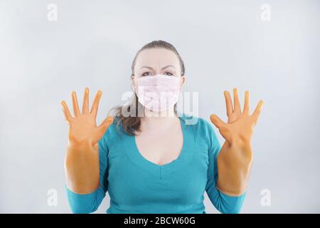 kaukasische Hausfrau mittleren Alters trägt eine schützende medizinische Maske und große Gummihandschuhe für Sterilität auf weißem Hintergrund, eine Frau in einem Haus-Pullover Stockfoto