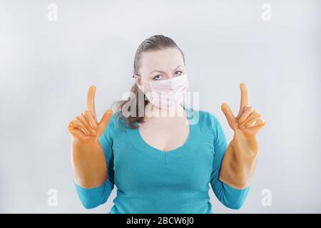 kaukasische Hausfrau mittleren Alters trägt eine schützende medizinische Maske und große Gummihandschuhe für Sterilität auf weißem Hintergrund, eine Frau in einem Haus-Pullover Stockfoto