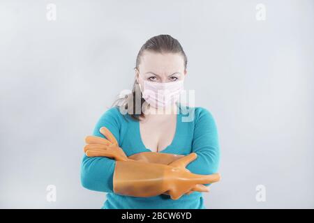 kaukasische Hausfrau mittleren Alters trägt eine schützende medizinische Maske und große Gummihandschuhe für Sterilität auf weißem Hintergrund, eine Frau in einem Haus-Pullover Stockfoto