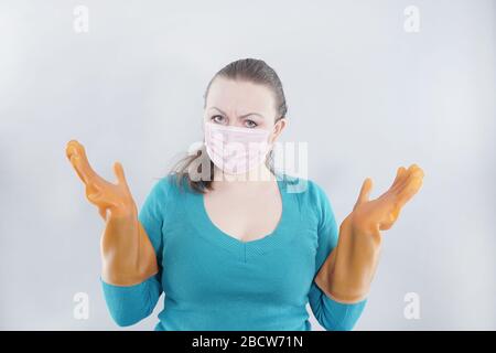 kaukasische Hausfrau mittleren Alters trägt eine schützende medizinische Maske und große Gummihandschuhe für Sterilität auf weißem Hintergrund, eine Frau in einem Haus-Pullover Stockfoto