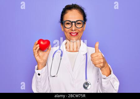Frau des leitenden Kardiologen im mittleren Alter, die rotes Herz über violettem Hintergrund hält, glücklich mit einem großen Lächeln, das ein gutes Zeichen tut, Daumen nach oben mit den Fingern, Erregung Stockfoto