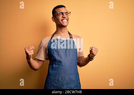 Junger gutaussehender afroamerikanischer Ladenbesitzer, der Schürze über gelbem Hintergrund trug, sehr glücklich und aufgeregt, indem er mit erhobenen Armen Gewinnergeste tat, schmilzt Stockfoto