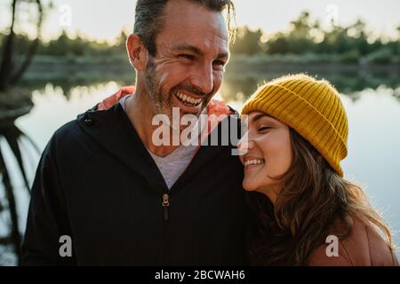 Liebendes Paar steht an einem See im Wald und lacht. Lächelnder Mann und Frau auf Campingausflug. Stockfoto