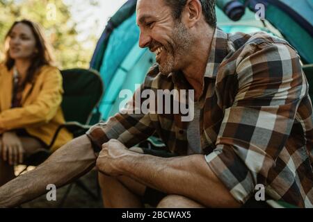 Reifer Mann, der mit einer Frau im Hintergrund auf dem Zeltplatz sitzt. Ein Paar auf dem Campen, das außerhalb des Zelts sitzt. Stockfoto