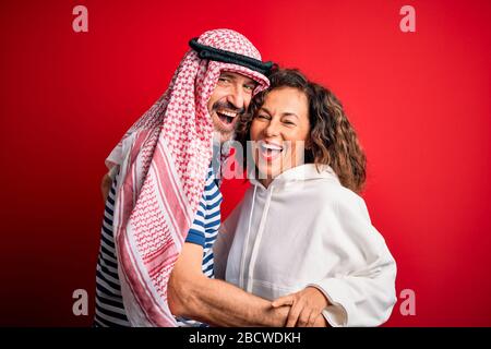 Schönes Paar im mittleren Alter mit arabischem Kopftuch und Sweatshirt, lächelnd glücklich und selbstbewusst. Mit einem Lächeln auf dem Gesicht stehend, umarmend über isoliertem Rot B. Stockfoto