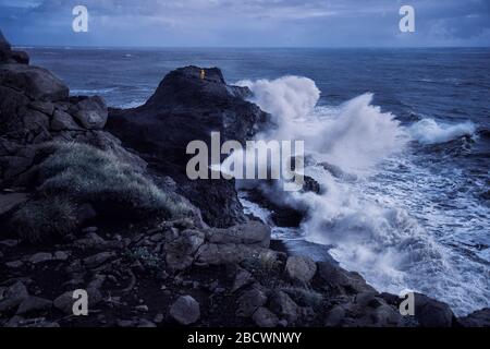 Mann in gelbem Regenmantel am Rand der Klippe mit riesigen Wellen, die am stürmischen Tag in Dyrholaey, Island, dagegen brechen Stockfoto