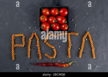 Italienisches Fusilli - Nudelwort. Rohe italienische Nudelfusilli mit Kirschtomaten in Bratpfanne und heißem Papier auf dunklem Hintergrund von oben. Flaches Lay Stockfoto