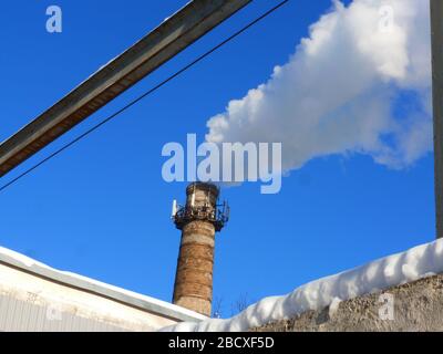 Rohre einer alten Fabrik werfen Wolken von giftigem weißem Rauch in den Himmel, die die Atmosphäre schädigen. Urbaner Smog aus Rauch aus Kesselhäusern. Weißer Rauch aus einem Kamin gegen einen blauen klaren Himmel. Stockfoto