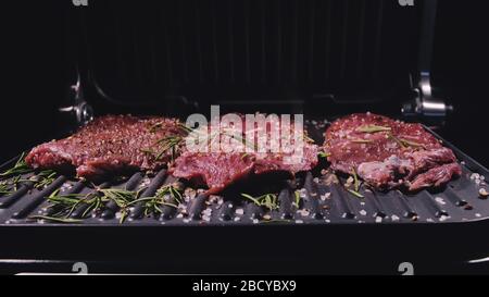 Köstliches, saftiges Steak-Kochen auf dem Grill. Braten Sie das Rindfleisch auf einem elektrischen Bräter, Rosmarin, schwarzem Pfeffer und Salz. Zeitlupe. Stockfoto