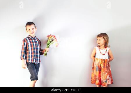 Der kleine Junge im Pflasterhemd gibt dem kleinen Mädchen im hellen Sommerkleid einen Blumenstrauß mit Tulpen. Stockfoto