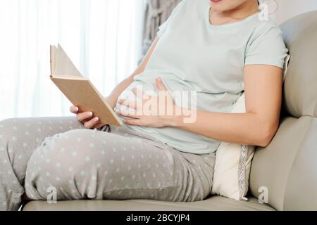 Junge schwangere Frau in Pyjamas, die ihren Bauch berühren, wenn sie ein Buch oder eine Zeitschrift liest Stockfoto