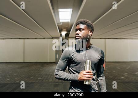 Porträt eines ernsthaften jungen schwarzen Sportlers, der Musik mit Ohrhörern und Trinkwasser hört Stockfoto