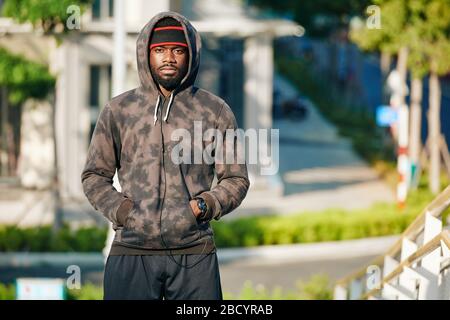 Seriöser junger Jogger im Hoodie, der die Hände in die Tasche auf dem Bauch steckt und die Kamera betrachtet Stockfoto