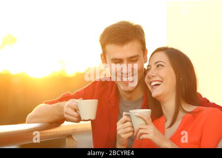 Ein glückliches Paar, das sich mit Kaffeetassen austresst und bei Sonnenuntergang auf einem Balkon zu Hause steht Stockfoto