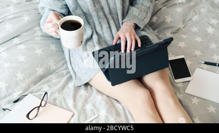 Junge Frau, die zu Hause am Tablet mit Tastatur arbeitet und Kaffee im Bett trinkt. Kuscheliger, hygge-Stil. Arbeiten zu Hause, sicher bleiben, Fernschulungskonzept Stockfoto