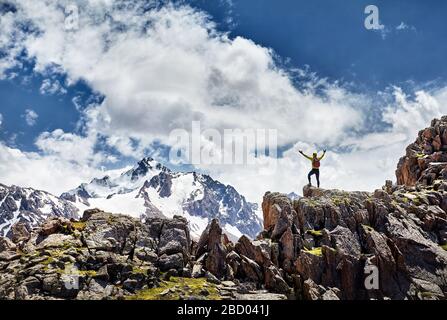 Wanderer in gelbem Hemd mit Rucksack, der auf dem Felsen steht, mit aufsteigenden Händen und Blick auf verschneite Berge bei bewölktem Himmel Stockfoto