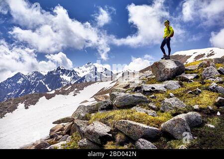 Wanderer in gelbem Hemd mit Rucksack, der auf dem Felsen steht und die Aussicht auf verschneite Berge bei bewölktem Himmel genießt Stockfoto