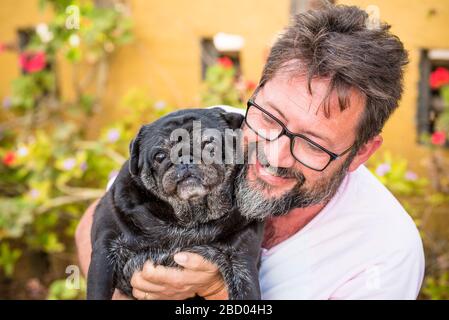 Im Freien schönes Porträt eines erwachsenen kaukasischen Mannes mit schwarzem Bart und gleicher Farbe alter lustiger Hundepug - Garten im Hintergrund und bestes Freundschafts-Liebeskonzept Stockfoto