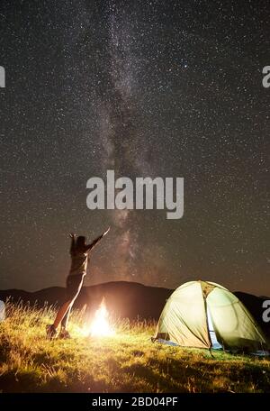 Fröhliche weibliche Wanderer, die sich in der Sommernacht beim Campen neben dem Lagerfeuer und dem leuchtenden Touristenzelt entspannen. Junge Frau hält die Hände hoch und genießt einen Blick auf den Nachthimmel voller Sterne und Milchstraße. Stockfoto