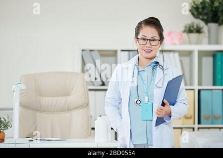Portrait eines positiv gereiften asiatischen Arztes mit Ordner, die in ihrem Büro stehen und mit der Kamera lächeln Stockfoto