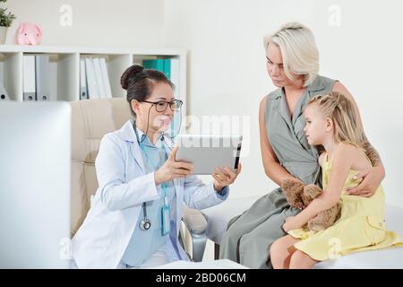 Reifer asiatischer Kinderarzt, der die Ergebnisse medizinischer Tests auf dem Bildschirm des Tablet-Computers der Mutter des kleinen Mädchens zeigt Stockfoto