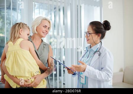 Lächelnder Kinderarzt mit Dokument in der Hand, der mit der Mutter des kleinen Mädchens über ihre Gesundheit spricht Stockfoto