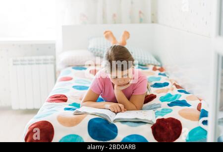 Süßes Tween Mädchen im rosafarbenen Lesebuch mit T-Shirt, das auf dem Bett liegt, im hellen Zimmer zu Hause Stockfoto