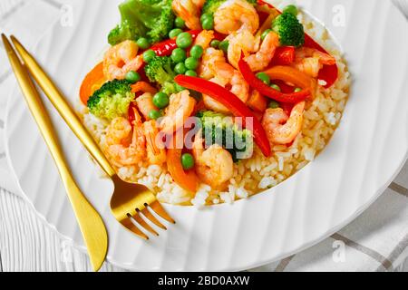 Nahaufnahme von gebratenen Garnelen mit Paprika, Zwiebel, Brokkoli und grünen Erbsen auf gedämpftem Reis auf einem weißen Teller auf einem Holztisch Stockfoto