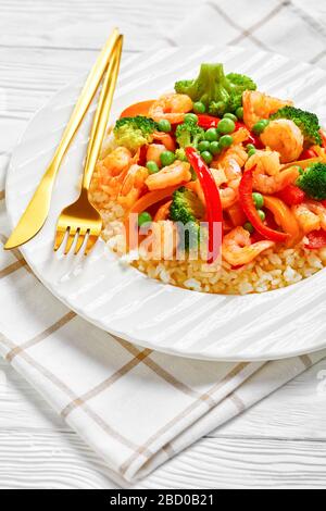 Nahaufnahme von gebratenen Garnelen mit Paprika, Zwiebel, Brokkoli und grünen Erbsen auf gedämpftem Reis auf einem weißen Teller auf einem Holztisch, vertikale Ansicht Stockfoto