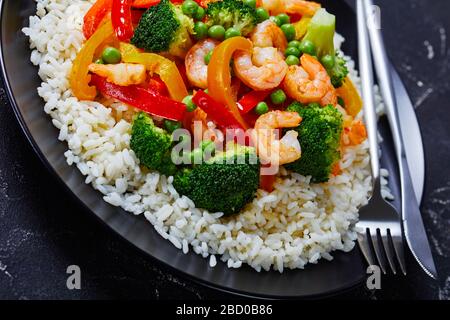 Nahaufnahme einer Portion gebratener Garnelen mit Paprika, Zwiebel, Brokkoli und grünem Erbsen auf gedämpftem Reis auf einem schwarzen Teller auf einem Betontisch Stockfoto