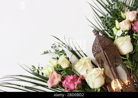 Marokkanische, arabische Laterne aus Vintage-Bronze mit glühender Kerze, grünen Zweigen, rosafarbenen Rosenblüten und Palmblättern auf weißem Tischhintergrund. Begrüßung Stockfoto