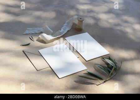 Leere Grußkarten, Umschlag, Seidenband und grüne olivgrüne Äste bei Sonnenlicht. Hintergrund aus goldenem Marmor mit Schatten. Sommerhochzeit Schreibwaren Stockfoto