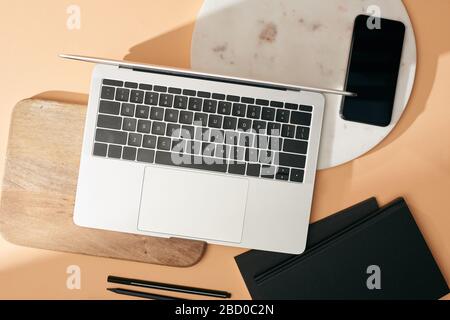 Draufsicht auf Laptop auf Holz- und Marmorplatten, Smartphone, Notizbücher und Stifte auf beigefarbenem Hintergrund Stockfoto